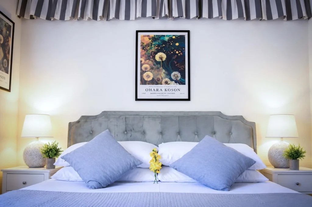 Letto matrimoniale con testata in velluto grigio, cuscini azzurri e quadri botanici alla Dimora di Arianna a Cariati.
