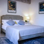 Letto matrimoniale con testata grigia, cuscini azzurri, quadri di Ohara Koson e lampade da comodino accese alla Dimora di Arianna a Cariati.