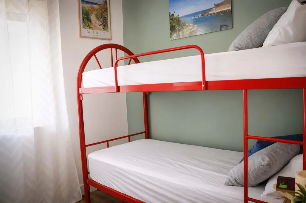 Letto a castello in metallo rosso con cuscini grigi e blu davanti a una parete verde salvia nella casa vacanze La Dimora di Arianna.
