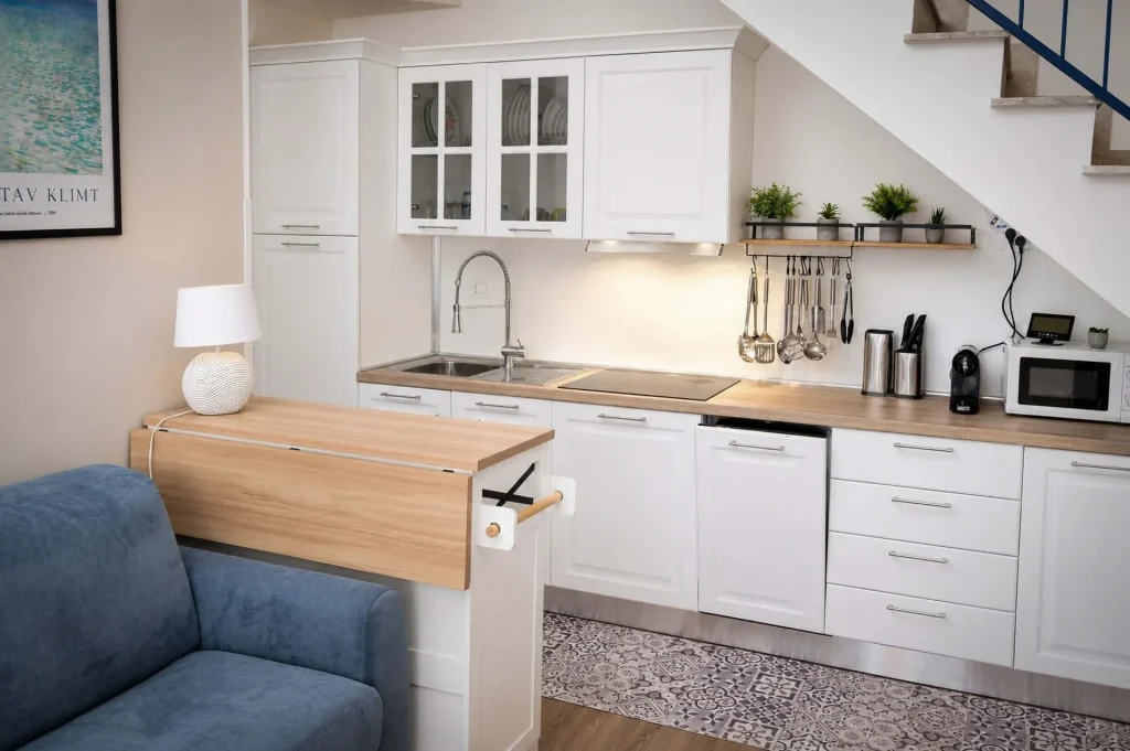Cucina moderna bianca ad angolo sotto la scala con piano cottura a induzione, lavastoviglie e divano azzurro in primo piano a Cariati.