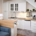 Cucina moderna bianca ad angolo sotto la scala con piano cottura a induzione, lavastoviglie e divano azzurro in primo piano a Cariati.