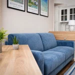 Soggiorno moderno con divano azzurro, tavolino in legno chiaro, lampada turchese e scorcio della cucina a Cariati Marina.