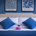 Letto matrimoniale con testata beige, cuscini blu cobalto e quadri di Henri Matisse su parete blu alla Dimora di Arianna a Cariati.