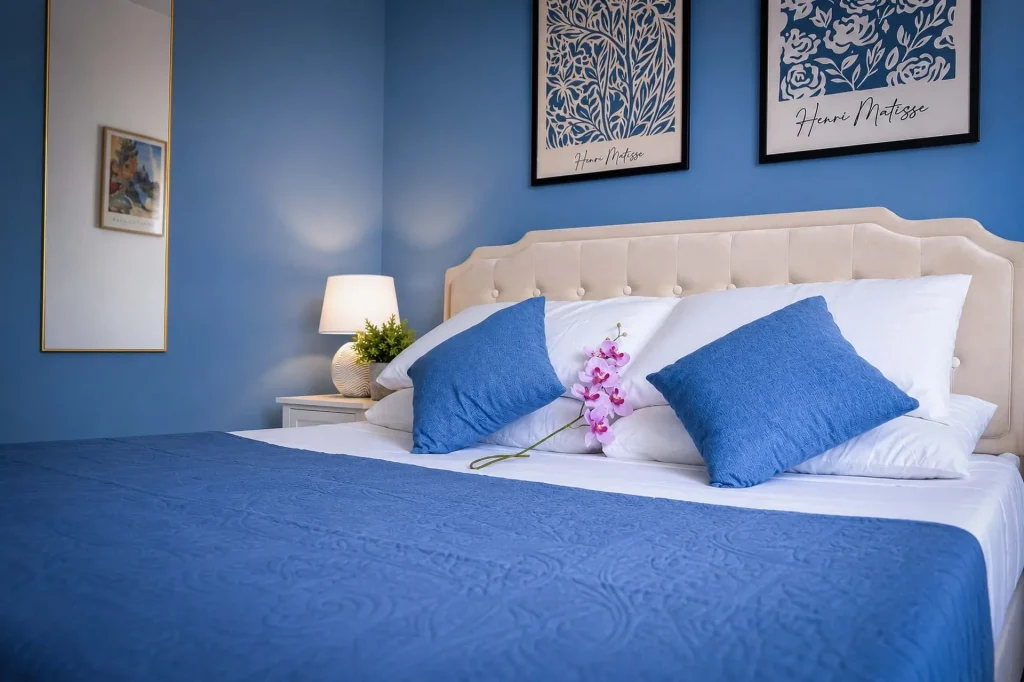 Vista angolare della seconda camera da letto con lenzuola bianche, copriletto blu, specchio a parete e quadri d'arte a Cariati.