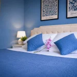 Vista angolare della seconda camera da letto con lenzuola bianche, copriletto blu, specchio a parete e quadri d'arte a Cariati.