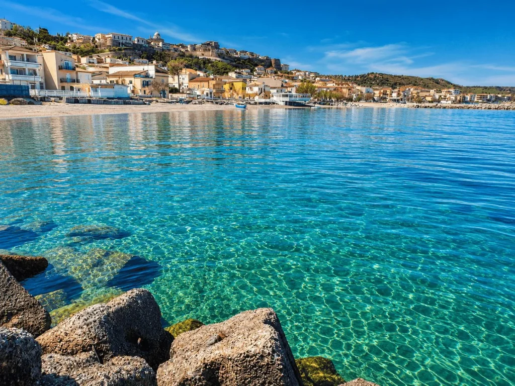 Mare cristallino turchese con scogli in primo piano e il borgo antico di Cariati Marina sullo sfondo in una giornata di sole.