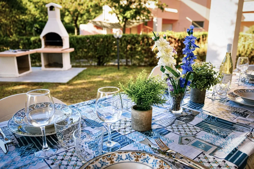Tavola apparecchiata all'esterno con piatti decorati in ceramica, bicchieri di vetro, fiori freschi e barbecue sullo sfondo a Cariati.