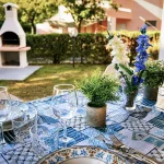 Tavola apparecchiata all'esterno con piatti decorati in ceramica, bicchieri di vetro, fiori freschi e barbecue sullo sfondo a Cariati.